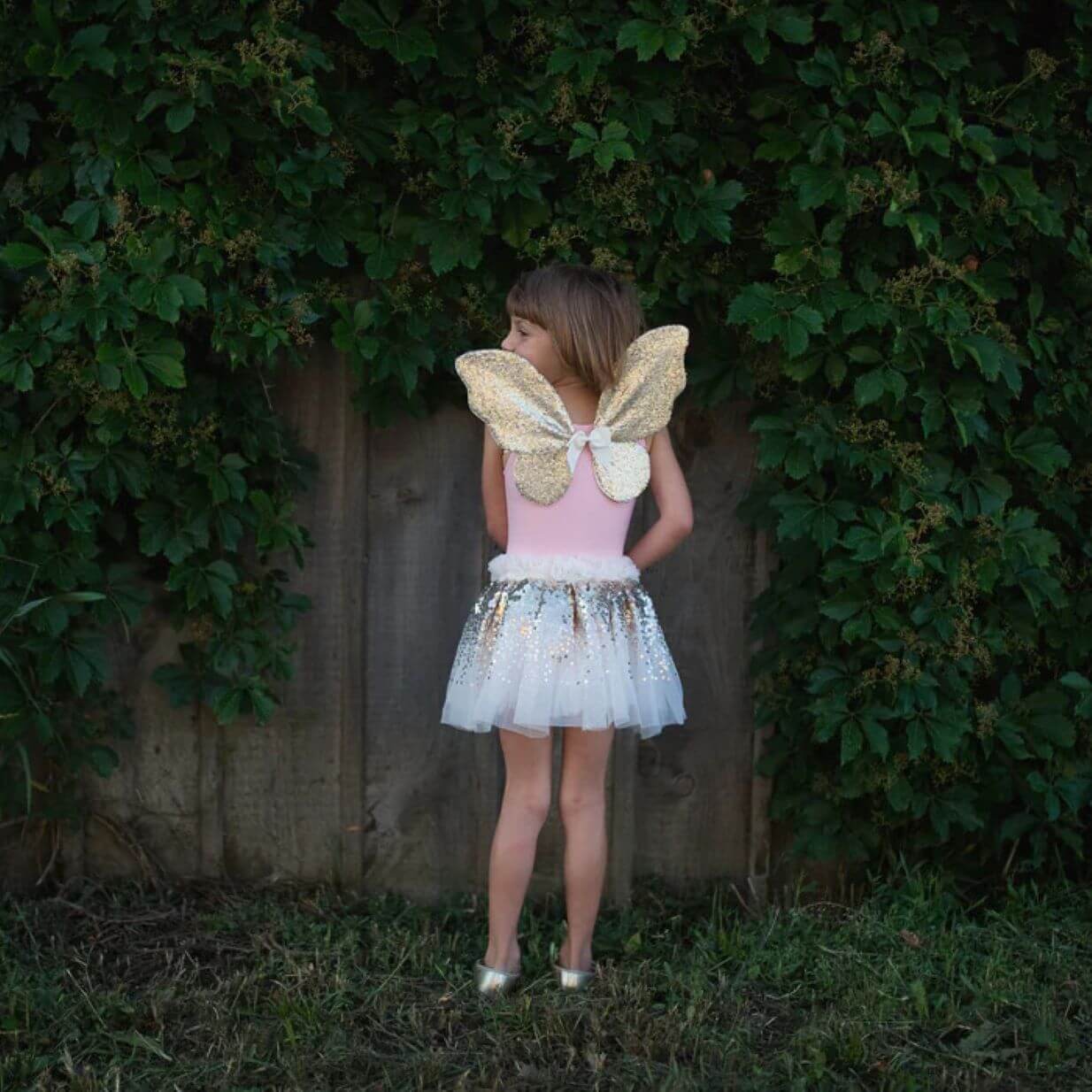 back view of young girl wearing pink top, gold sequin fairy wings and white tutu skirt with gold sequins