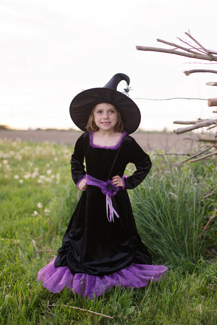 A young girl wearing a black and purple witch costume, including a pointed black hat, black dress with purple trim, and a purple tulle skirt, standing in a grassy outdoor field.