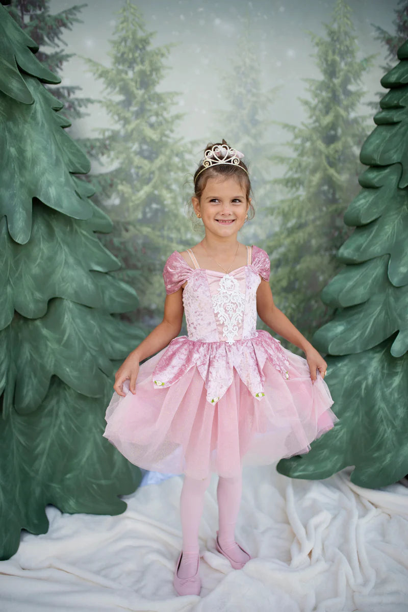 Young girl in a pink dress with a tiara standing in front of green trees.