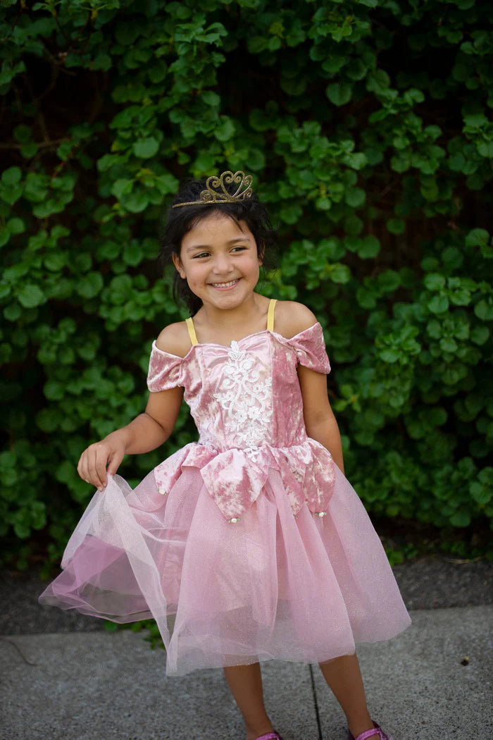 Young girl in a pink dress with a tiara standing in front of green foliage.