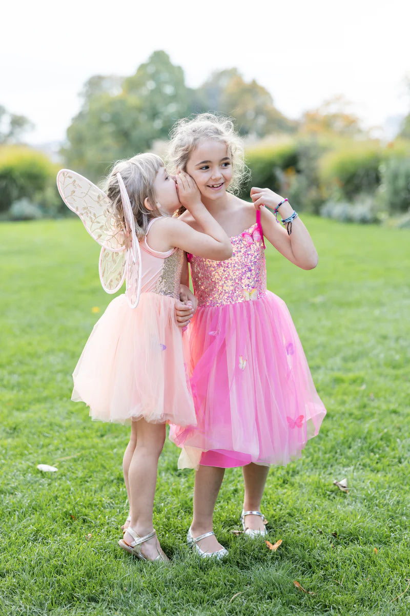 Two young girls in pink dresses standing on grass, one with fairy wings.