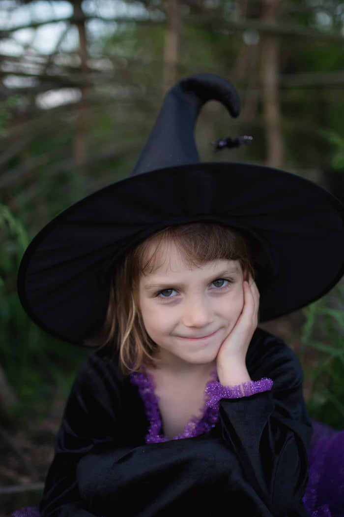 A young girl wearing a black witch costume with a pointed black hat and purple trim, posing outdoors in a forest setting.