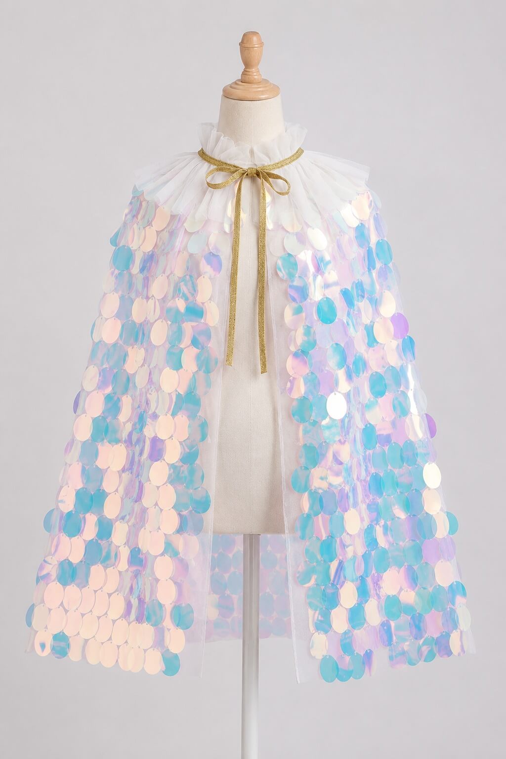 A white tulle cape with iridescent pastel sequins in shades of blue, pink, and purple, tied with a gold ribbon at the collar, displayed on a mannequin against a light gray background.