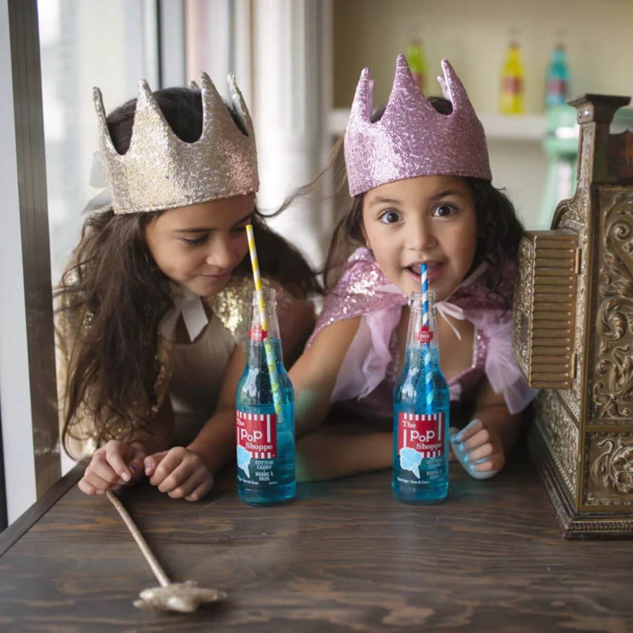 two young girls drinking fizzy drinks from a bottle with a gold sequin fabric crown and a pink sequin fabric crown