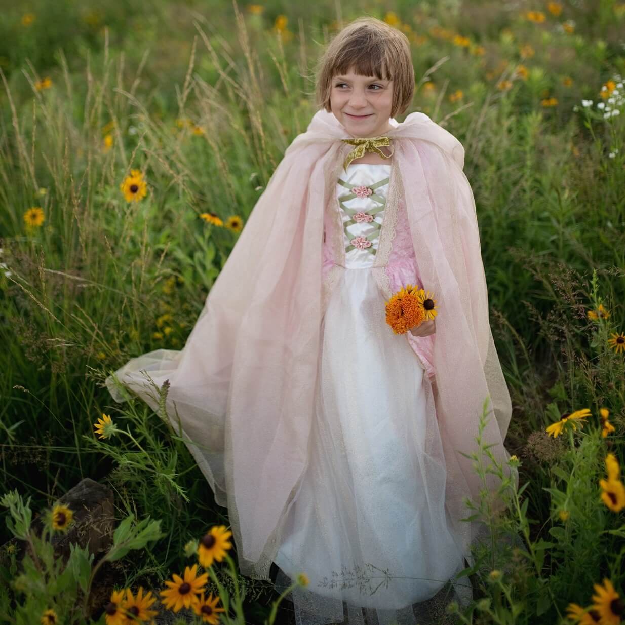 Girl wearing pink and gold princess cape in a field holding an orange flower