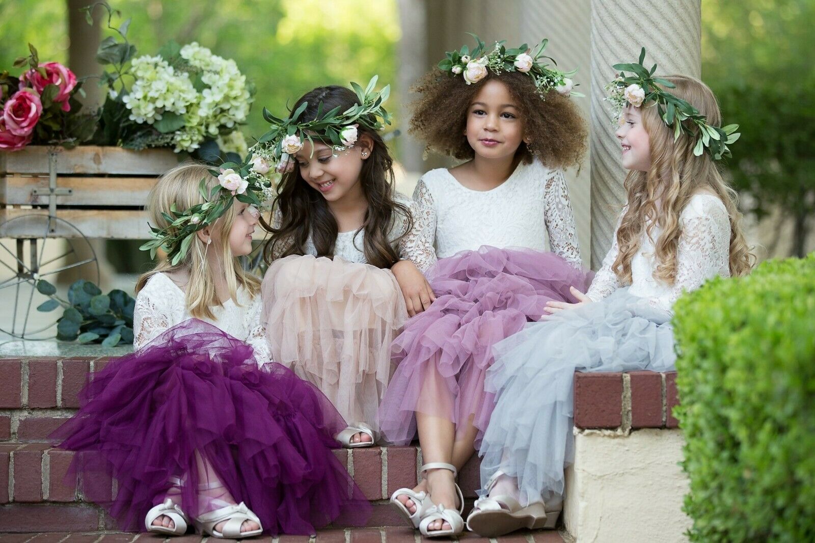 group of 4 girls sitting in dresses with various coloured ruffle skirts, with natural green foliage and rose head garlands